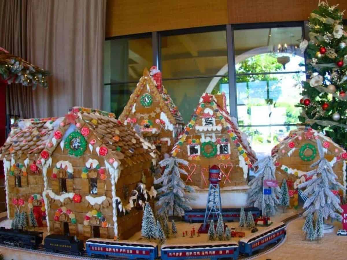 A picturesque gingerbread village display featuring several detailed houses decorated with icing, candy canes, and gumdrops. The setup includes miniature snow-covered trees and a toy train encircling the scene, all positioned in a brightly lit indoor space with a Christmas tree in the background.