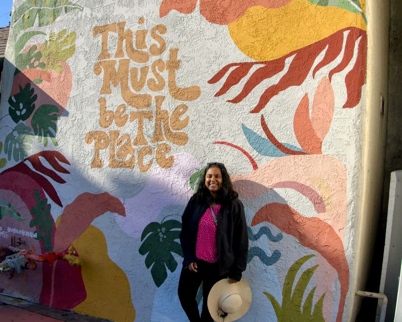 A colorful mural on a wall with the text "This Must Be the Place" surrounded by tropical leaf patterns and vibrant hues. Shreeyeh stands smiling in front of the mural, holding a wide-brimmed hat.