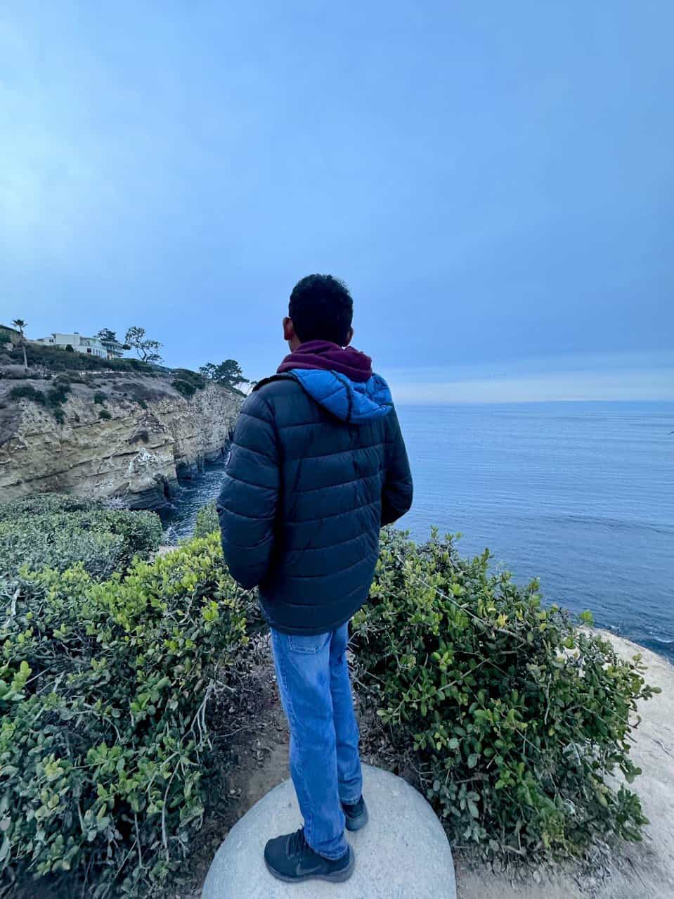 Kiran in a black jacket and jeans stands on a rock overlooking the ocean from a cliff in La Jolla, surrounded by green shrubs under a cloudy blue sky.