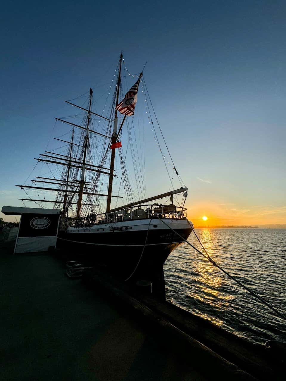 The "Star of India" ship docked at the waterfront, silhouetted against a vivid orange sunset, with calm waters reflecting the warm hues and an American flag flying atop the masts.