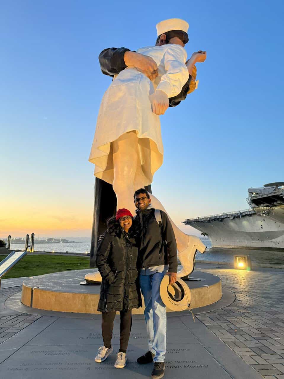 Shreeyeh and Kiran pose in front of the "Unconditional Surrender" statue depicting a sailor kissing a nurse, with the San Diego harbor and a naval ship visible in the background at sunset.