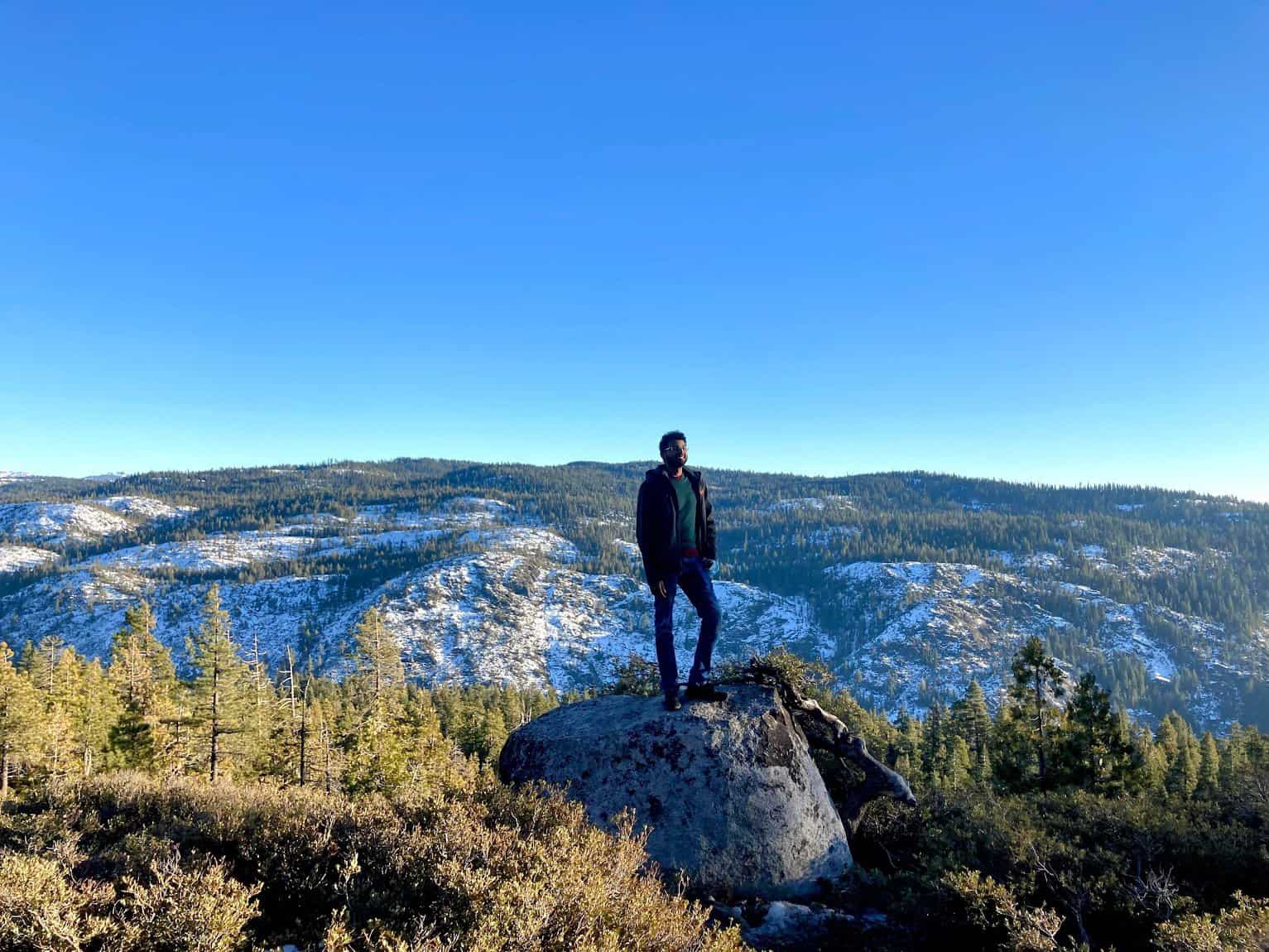 Kiran stands confidently on a large rock overlooking a stunning snowy mountain landscape. Pine trees and snow-dusted hills stretch out beneath a clear blue sky, capturing the serene beauty of nature.