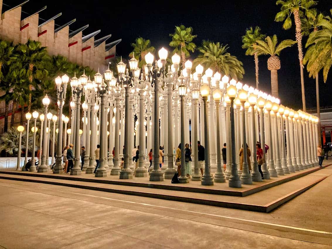 A nighttime view of the Urban Lights installation at the Los Angeles County Museum of Art (LACMA), featuring rows of glowing white street lamps surrounded by visitors.