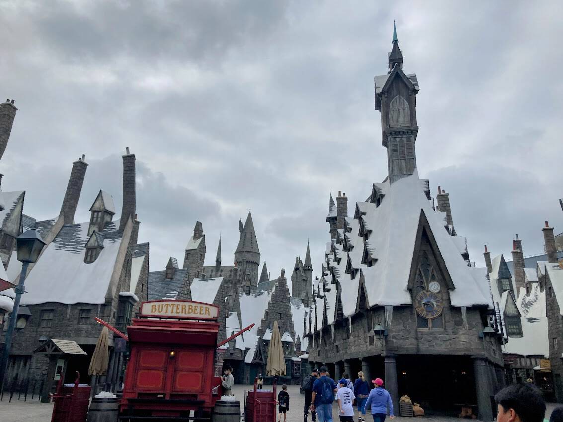 A view of the snow-covered rooftops and medieval-style architecture of Hogsmeade Village, with a red Butterbeer cart and visitors exploring the area.