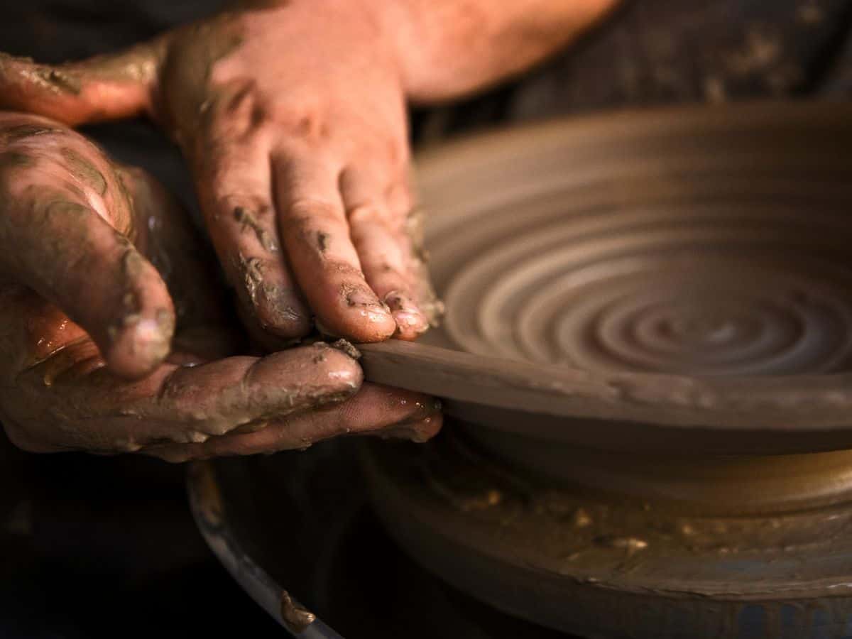 A close-up image of a person's hands covered in wet clay, skillfully shaping a spinning piece of pottery on a wheel. The circular grooves of the pottery highlight the creative process in progress.