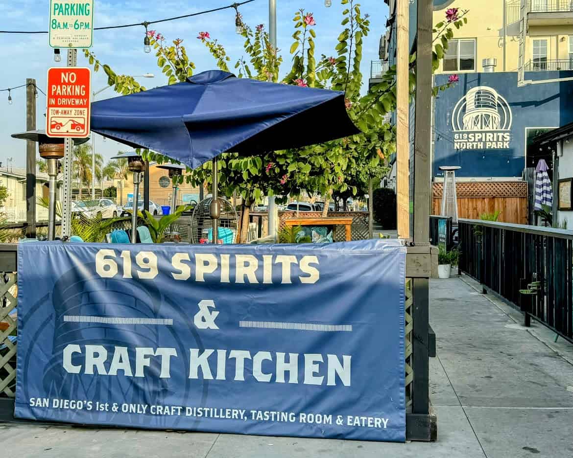 The patio of 619 Spirits & Craft Kitchen in North Park, with a banner reading "San Diego's 1st & Only Craft Distillery." Blue umbrellas, outdoor seating, and greenery create a cozy dining space.