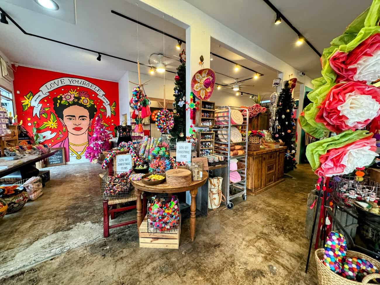 The interior of Artelexia, a colorful store featuring Mexican-inspired gifts and decor. A bold mural of Frida Kahlo on a red wall reads “Love Yourself” amidst a festive display of vibrant crafts and ornaments.