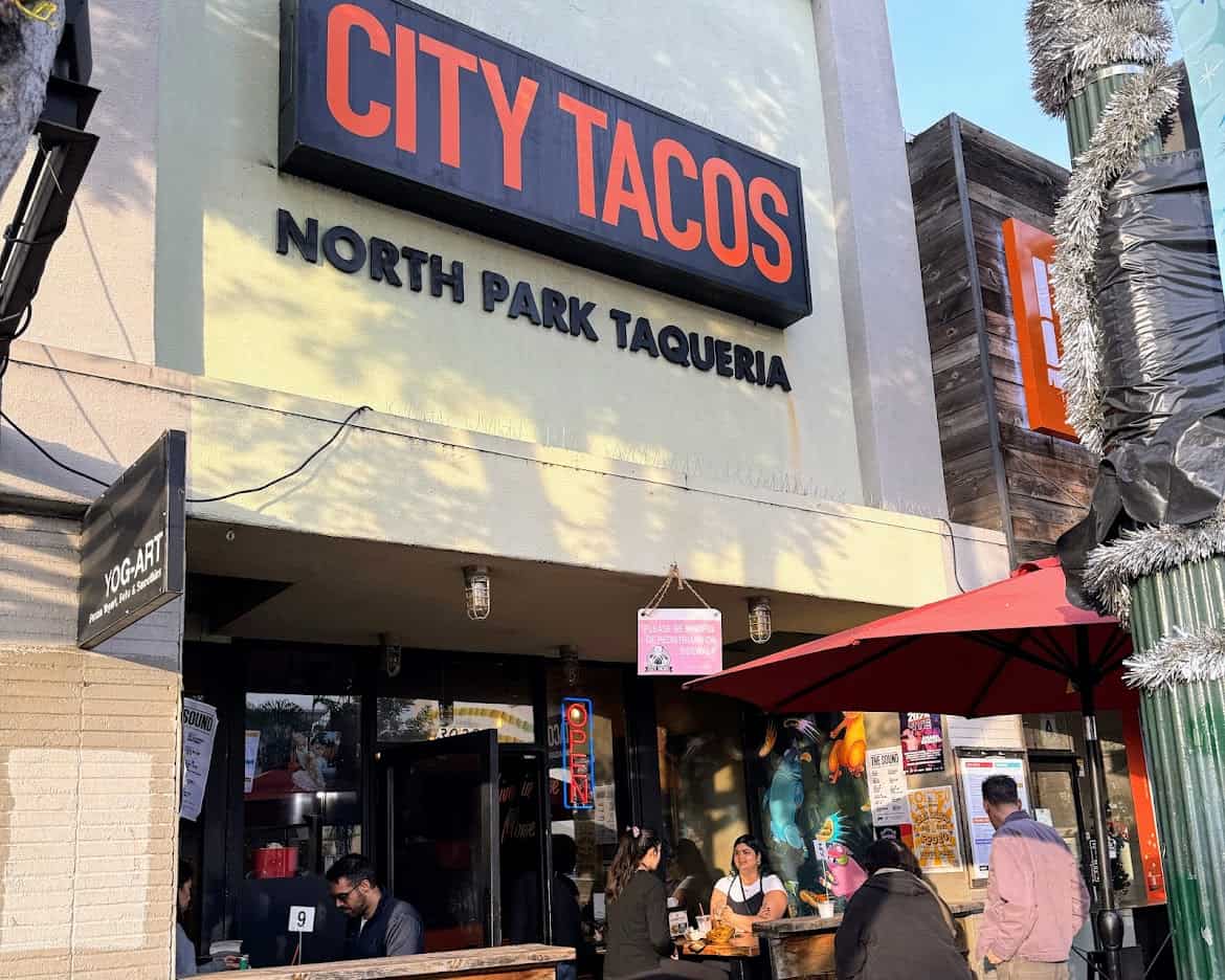 The exterior of City Tacos North Park Taqueria, a bustling taco shop with outdoor seating under a red umbrella. A neon “OPEN” sign and diners enjoying their food add to the lively atmosphere.