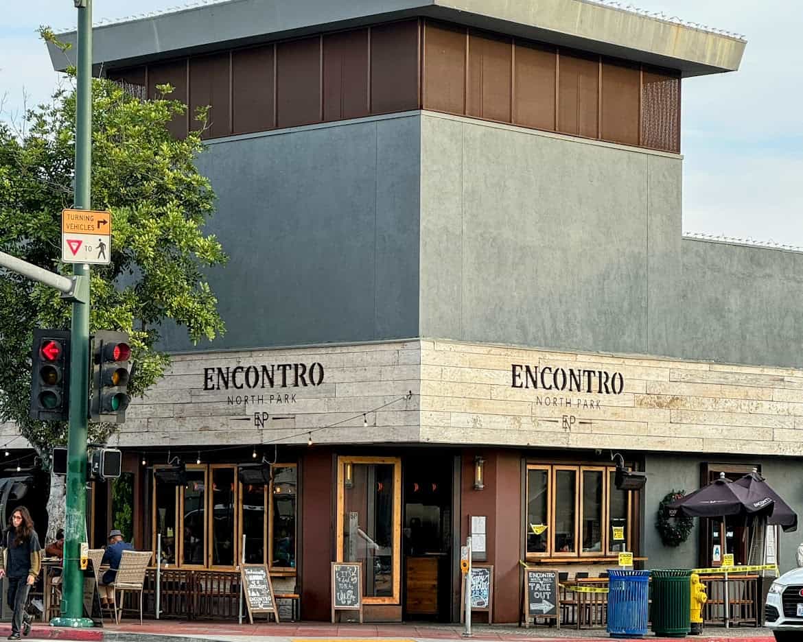 A corner view of Encontro North Park, a modern eatery with wooden accents and large windows. The building is surrounded by trees and a small outdoor seating area, with a pedestrian crossing and traffic light in the foreground.