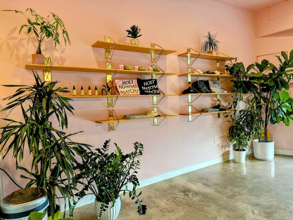 A serene corner of Holy Matcha, a tea cafe with pale pink walls, minimalist wooden shelves displaying matcha-themed items, and lush green plants in white pots for decoration.