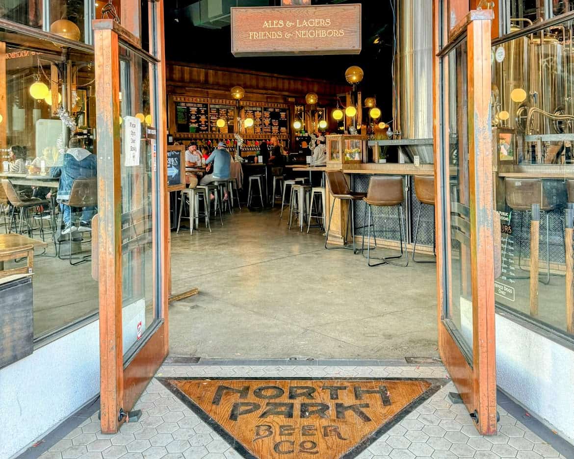 The entrance to North Park Beer Co., a brewery with an inviting interior featuring warm lighting and wooden decor. The open doors lead to a lively bar area with patrons enjoying drinks.