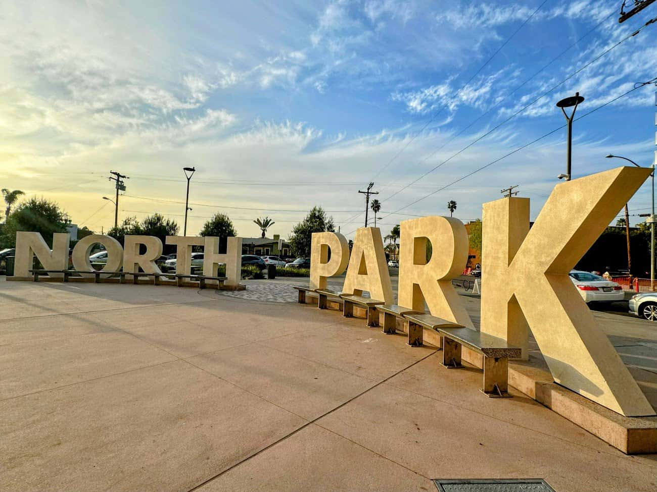 Large block letters spelling “NORTH PARK” at the Mini Park plaza, illuminated by the warm glow of the evening sun. The surrounding space features benches, greenery, and a paved walkway.
