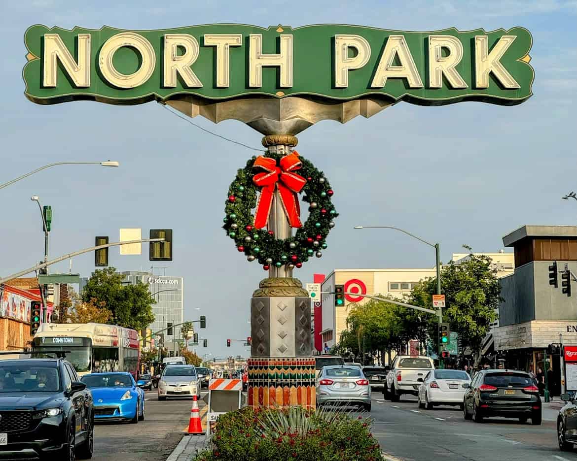 The iconic North Park sign in San Diego, adorned with a festive wreath and a red bow. The street below is busy with cars and lined with shops and buildings.