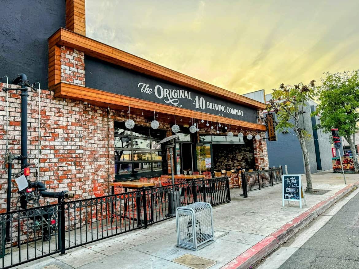 A photo of the Original 40 Brewing Company in North Park, San Diego, featuring a brick facade with a wooden trim and a black sign reading “The Original 40 Brewing Company.” Outdoor seating is enclosed by a black metal fence, and the sky has a golden evening hue.