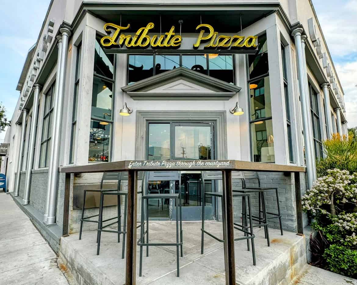 A stylish exterior of Tribute Pizza in North Park, San Diego, with a modern design, large windows, and a neon yellow sign that reads "Tribute Pizza." A small elevated table and chairs sit outside near the entrance, which has a sign indicating entry through the courtyard.