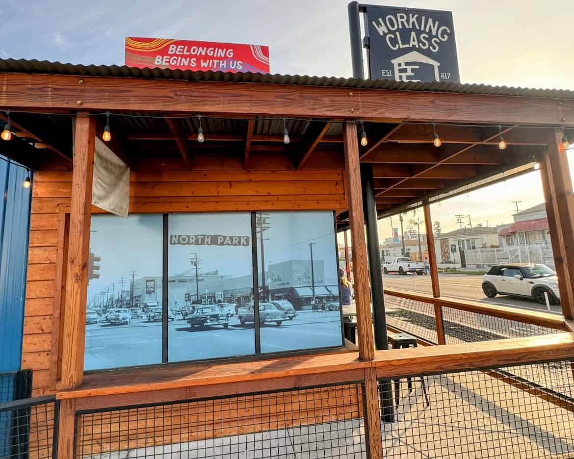 A wooden patio area of Working Class restaurant in North Park, showcasing a vintage-style photo of North Park and a festive sign reading “Belonging Begins With Us.” String lights and open seating add a welcoming vibe.