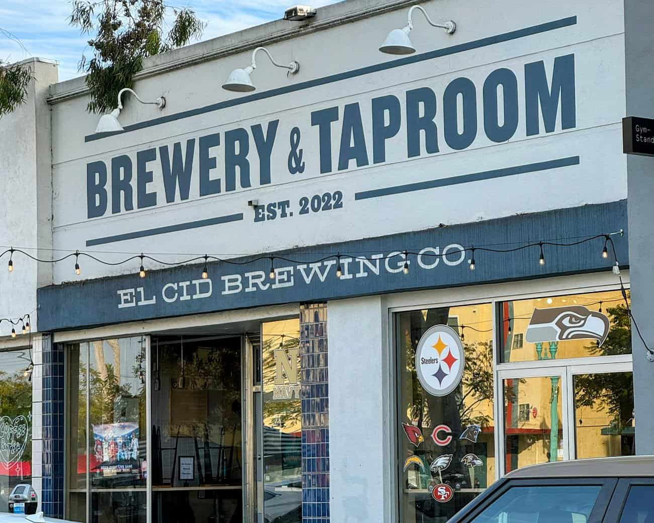 The entrance to El Cid Brewing Co., labeled as a "Brewery & Taproom" established in 2022, with string lights, a simple white facade, and sports-themed decor in the windows.