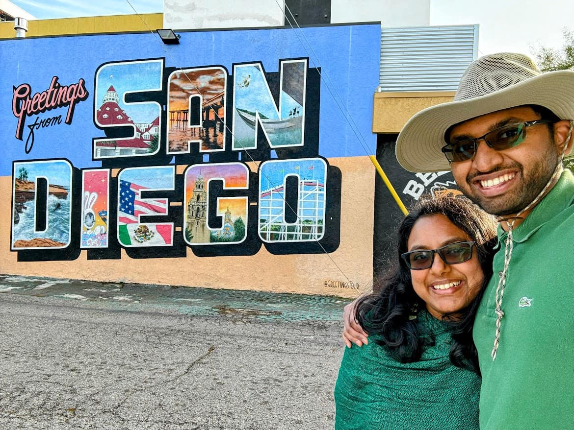 Kiran and Shreeyeh pose in front of the "Greetings from San Diego" mural in North Park, featuring colorful postcard-style letters showcasing iconic San Diego landmarks and imagery, like the Coronado Hotel, a beach pier, and local wildlife.