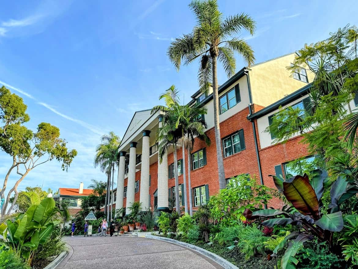 The Lafayette Hotel in North Park, San Diego, surrounded by lush greenery and tall palm trees. The red-brick building features large white columns and classic windows, with a welcoming walkway leading to the entrance.