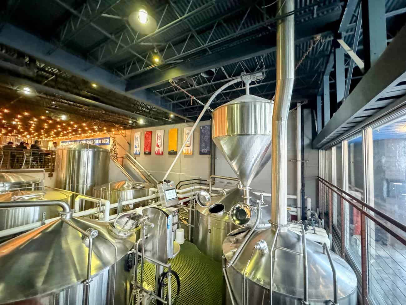 Inside Mike Hess Brewing in North Park, showcasing shiny stainless steel brewing tanks and equipment used in the beer-making process. The industrial-style ceiling is adorned with string lights, and colorful brewery banners decorate the wall in the background, creating a lively and modern atmosphere.