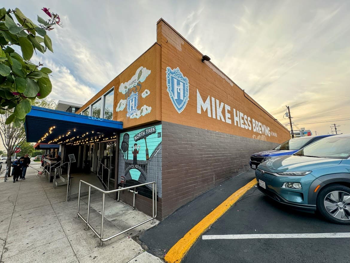 The orange brick building of Mike Hess Brewing in North Park, featuring a mural with the brewery’s logo and a shaded outdoor patio area lined with string lights.