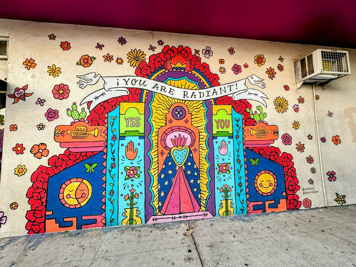 A colorful and uplifting mural with the text “You Are Radiant!” surrounded by flowers, bright patterns, and cheerful symbols like doves and musical notes on a vibrant wall.