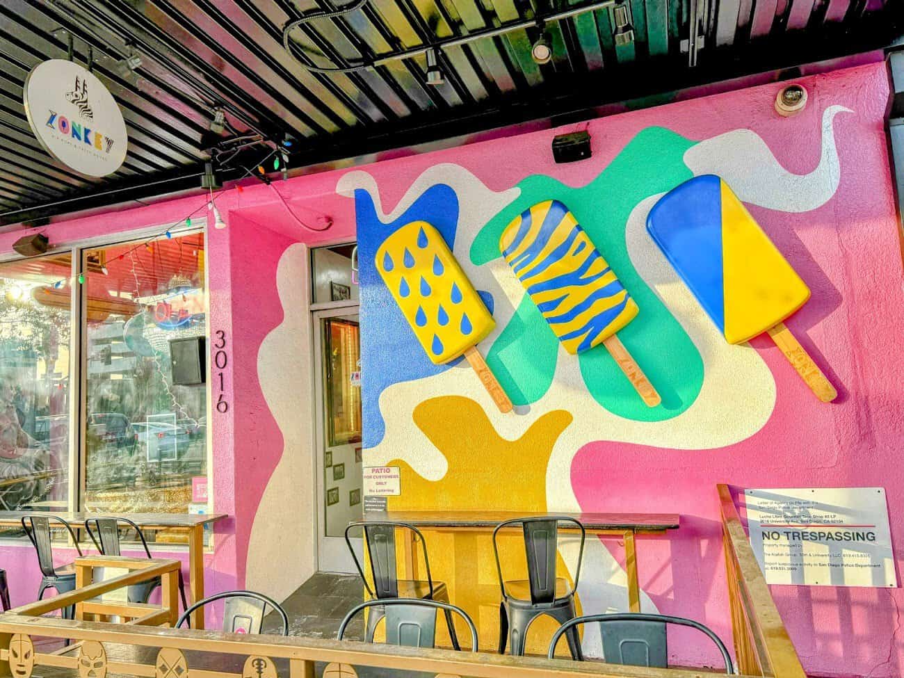 A vibrant storefront of Zonkey in North Park, with a playful mural of oversized popsicles painted in bold colors and patterns on a pink background. Outdoor seating with black chairs and wooden tables lines the entrance.