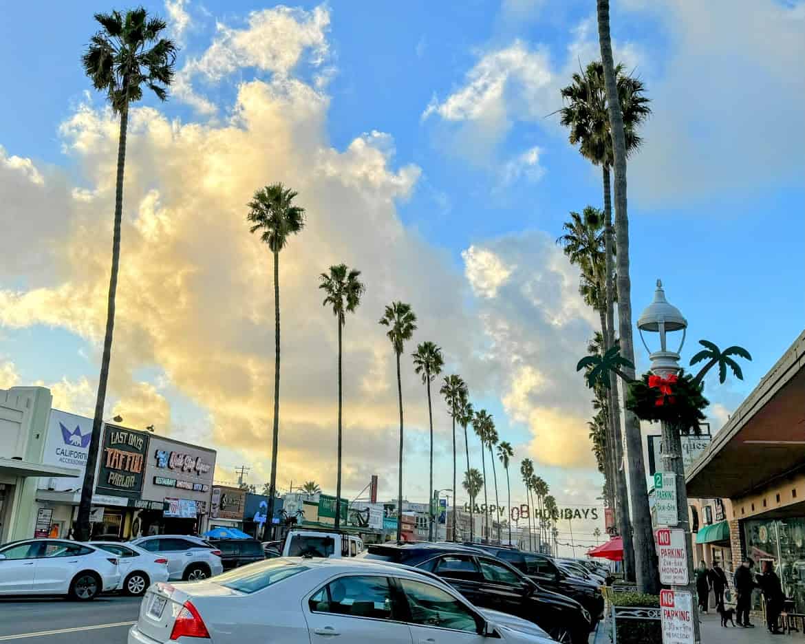 A bustling scene on Newport Avenue in Ocean Beach, with iconic palm trees lining the street, festive holiday decorations, and lively shops and cars filling the vibrant avenue.