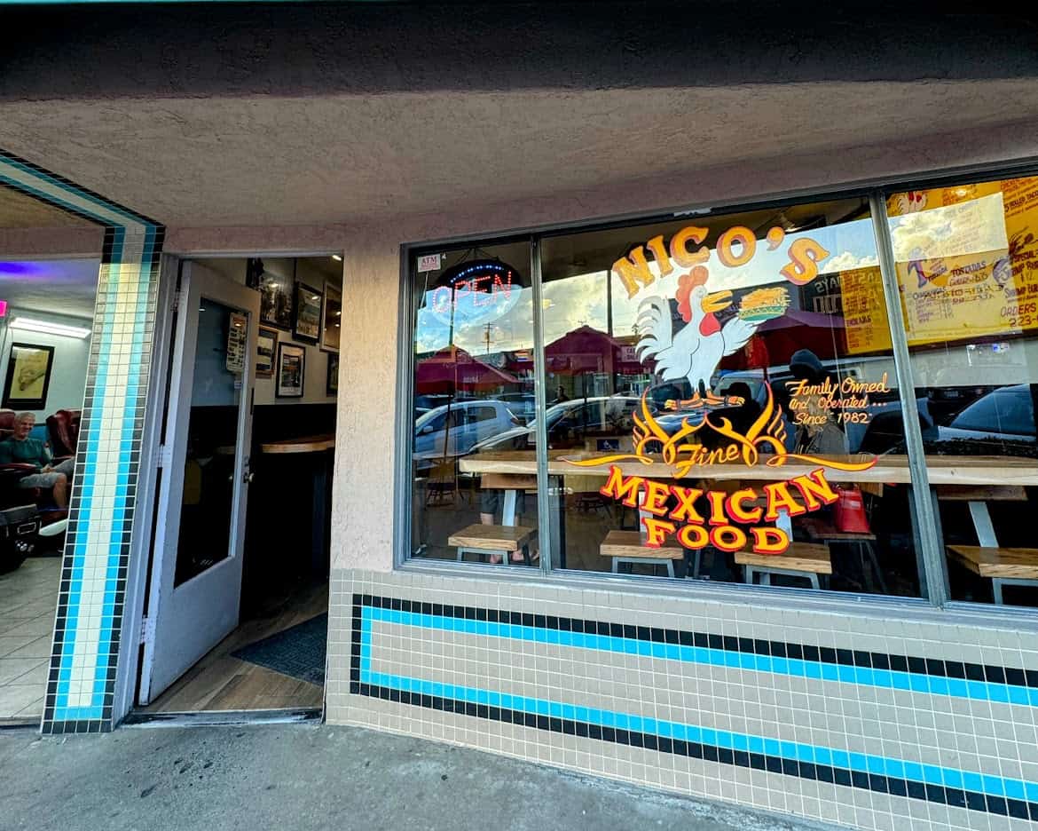 The entrance to Nico's Mexican Food in Ocean Beach, featuring a colorful window design with a rooster holding a taco and the text "Family Owned and Operated Since 1982." The inviting interior shows wooden tables and framed photos on the wall.
