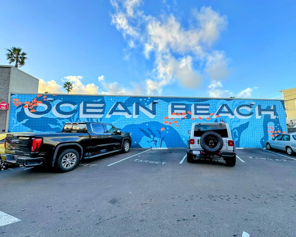 A vibrant mural in Ocean Beach featuring bold white text that reads "OCEAN BEACH" against a blue background with marine-themed illustrations, including fish, waves, and underwater shapes. The mural is located on a parking lot wall, with several vehicles parked in front of it.