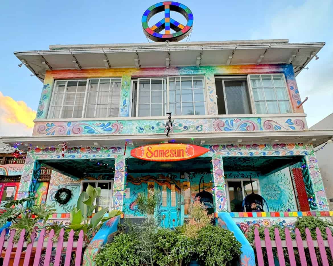 A colorful and artistic hostel in Ocean Beach, Samesun is painted with vibrant rainbow patterns and features a large peace sign on its roof. The front yard is decorated with plants, a pink picket fence, and festive lights.