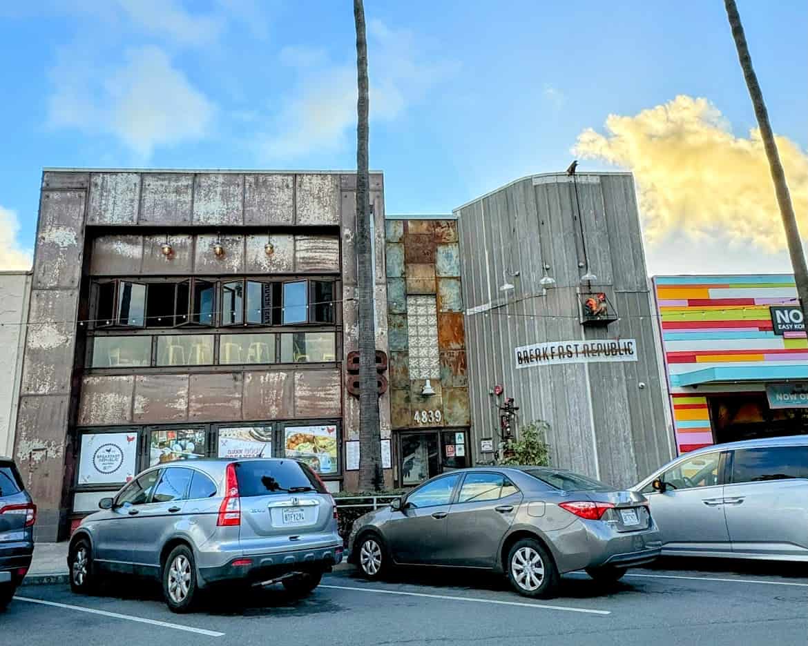 The rustic facade of Breakfast Republic in Ocean Beach, featuring a weathered wood and metal design. The quirky exterior contrasts with the vibrant neighboring buildings, including a colorful striped storefront.