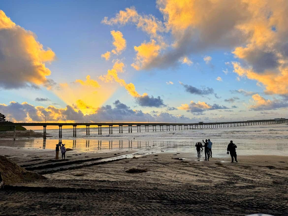 Ocean Beach Pier at sunset, with warm orange and yellow hues lighting up the clouds and silhouetted visitors enjoying the serene evening.