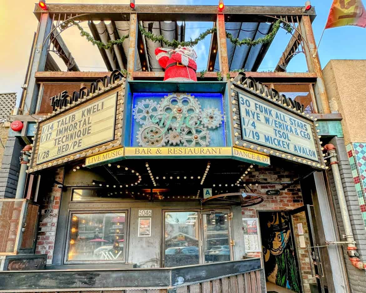 The Holding Company, a live music bar and restaurant in Ocean Beach, featuring a marquee advertising upcoming events. The industrial-style facade is adorned with holiday decorations and a small Santa figure on top.