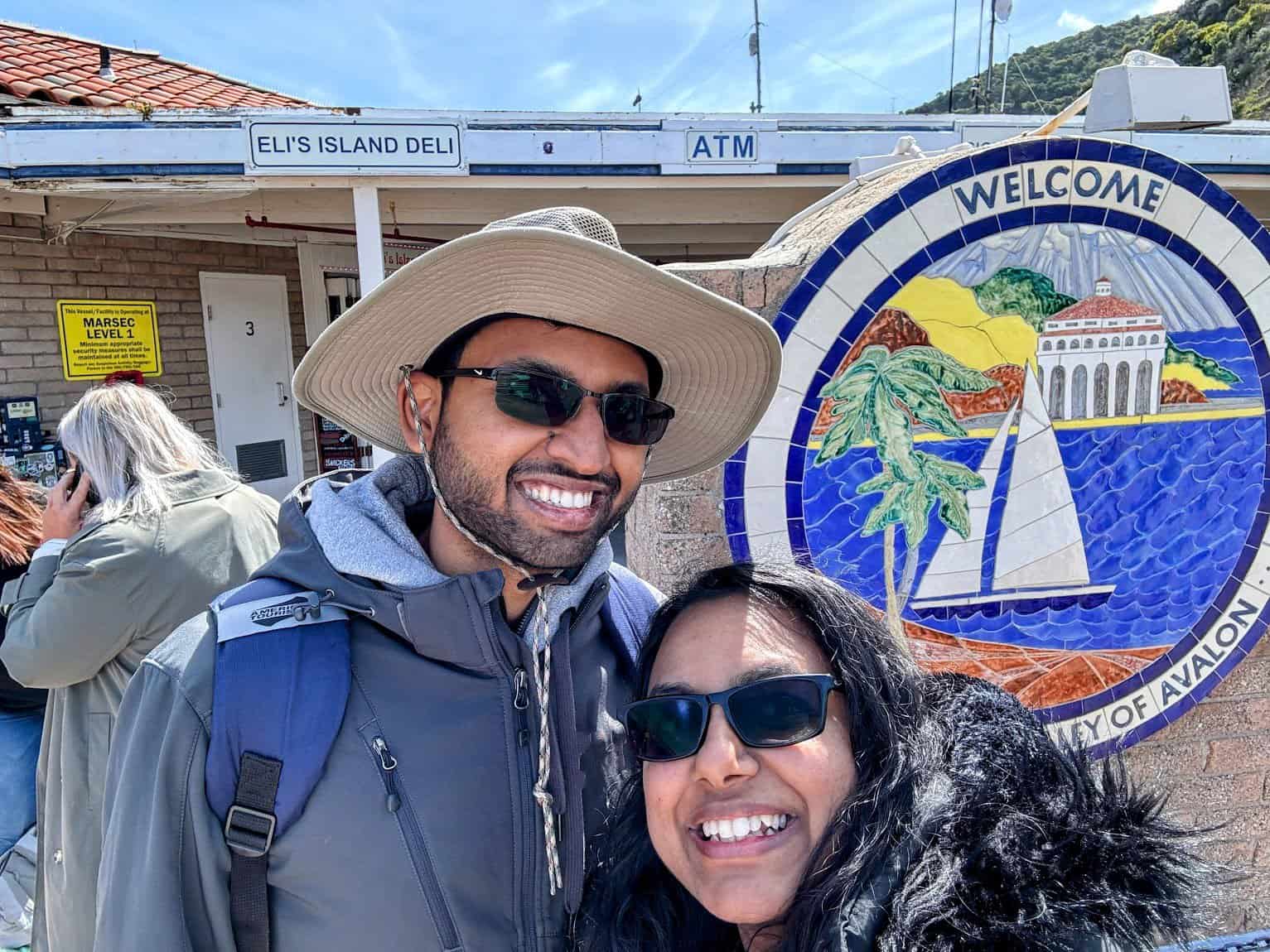 KIran and Shreeyeh posing in front of a colorful mosaic sign welcoming visitors to the City of Avalon, Catalina Island. The sign features imagery of a sailboat, palm trees, and the Avalon Casino against a vibrant backdrop, with Eli's Island Deli visible in the background.