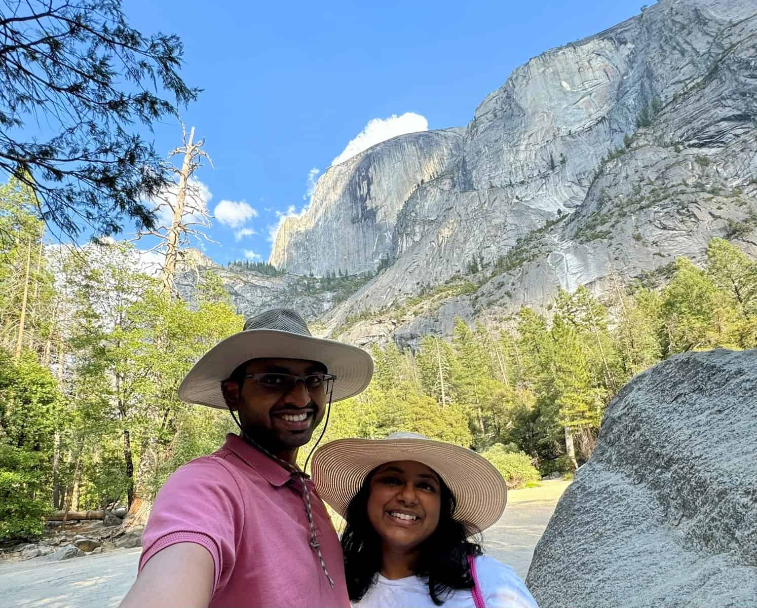 Kiran and Shreeyeh wearing wide-brimmed hats takes a selfie in front of towering granite cliffs and lush green trees at Yosemite National Park. The clear blue sky and dramatic rock formations create a stunning backdrop.