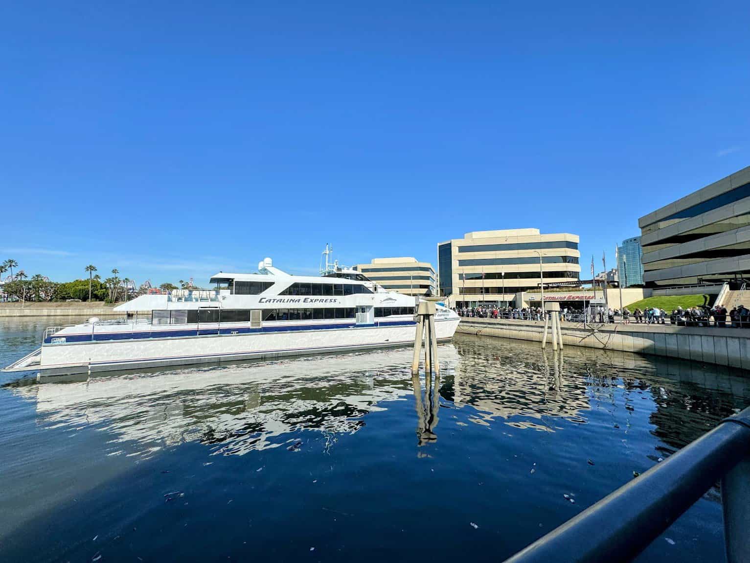 The Catalina Express ferry docked near the Long Beach harbor, with modern office buildings and clear blue skies in the background. Passengers are seen lining up near the boarding area.