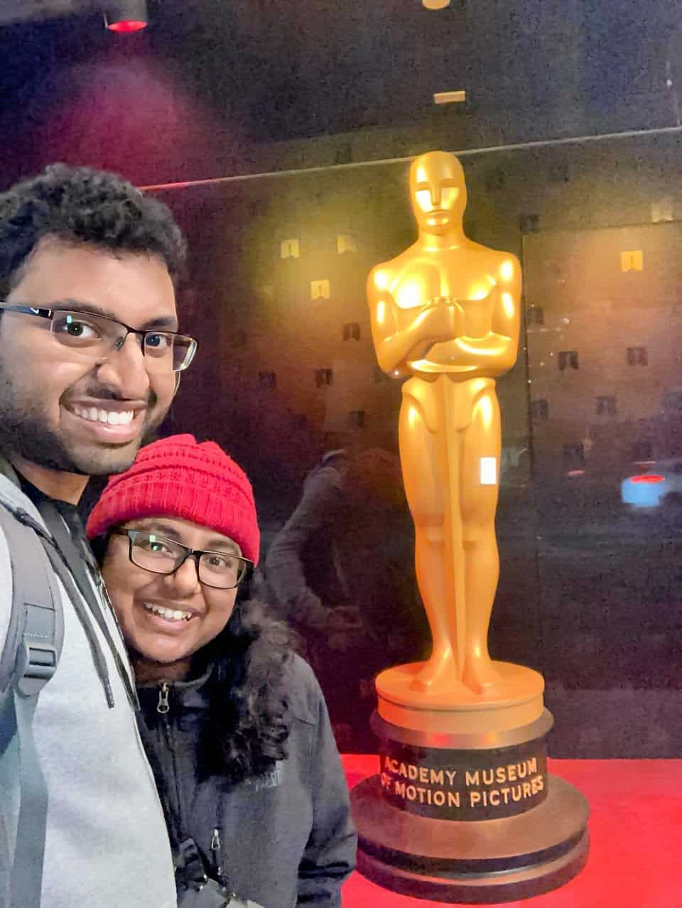 Kiran and Shreeyeh take a selfie in front of a large golden Oscar statue displayed at the Academy Museum of Motion Pictures. The exhibit is set against a dramatic, dark backdrop with subtle lighting highlighting the iconic award.