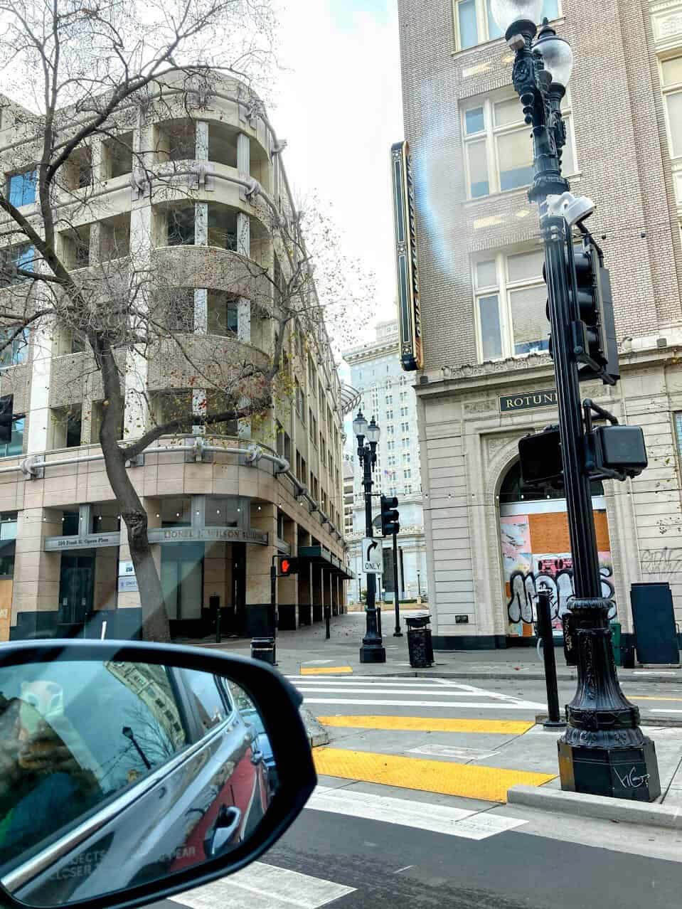 A street corner in Oakland with historic buildings, a lamp post, and crosswalks. The reflection of the street and buildings is visible in the side mirror of a car, adding a dynamic perspective to the urban scene.