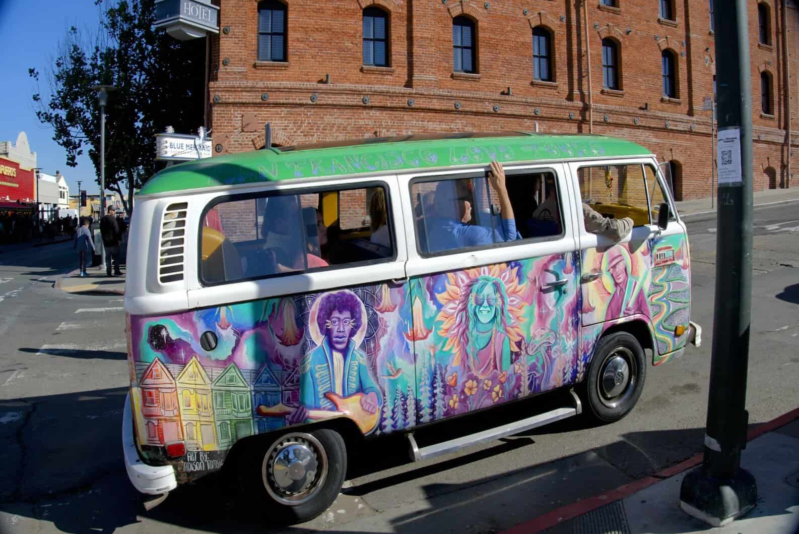A colorful retro Volkswagen bus decorated with vibrant murals featuring iconic San Francisco landmarks and figures, including painted houses and psychedelic art. The bus, marked "San Francisco Love Tours," is parked near a brick building in a lively urban setting.
