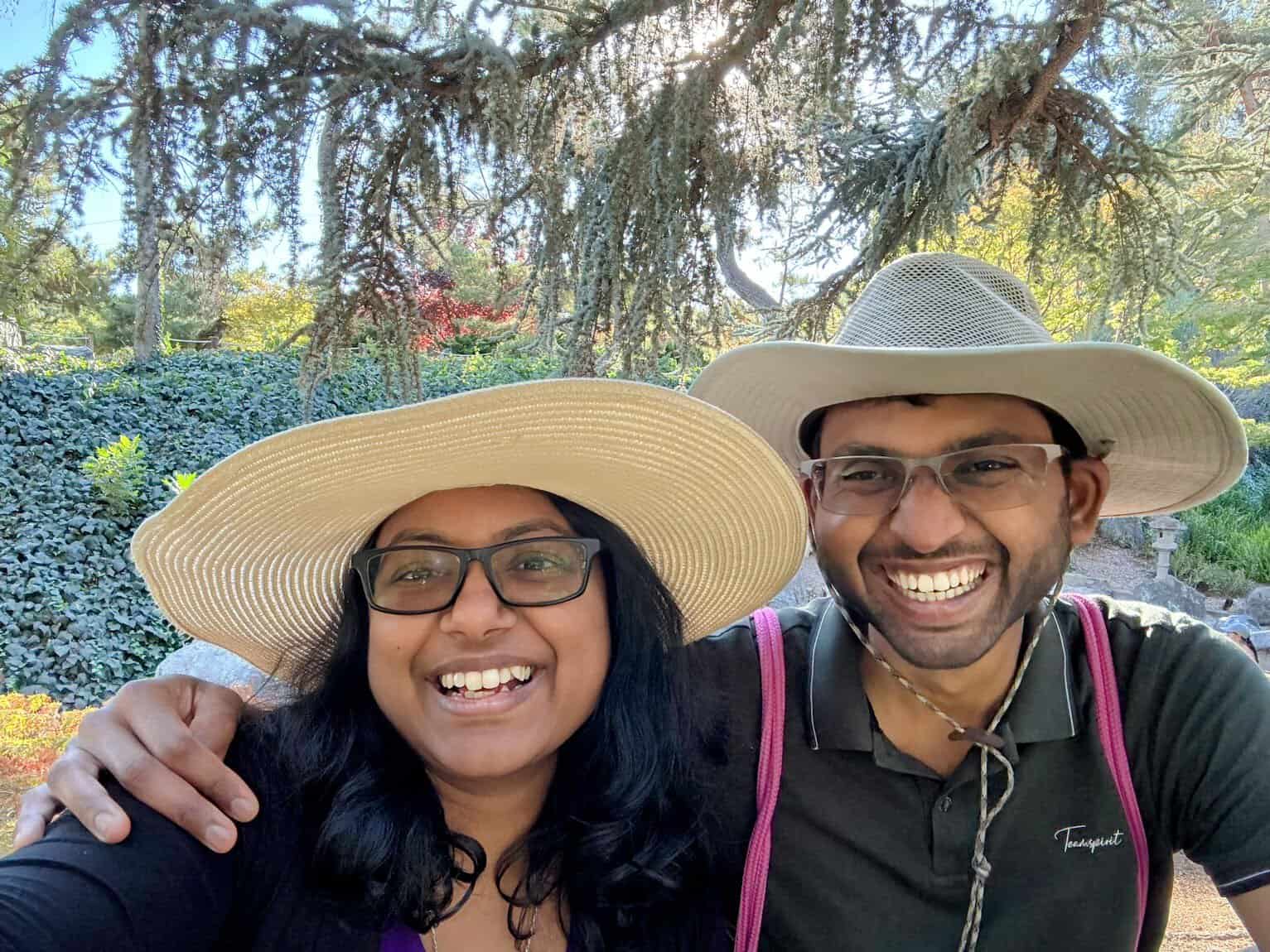 Shreeyeh and Kiran wearing sun hats smiles broadly for a selfie in a lush garden setting. The background features vibrant greenery, colorful foliage, and hanging tree branches creating a serene, natural ambiance.