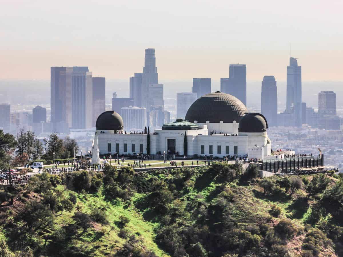 A panoramic view of Griffith Observatory in Los Angeles, with the skyscrapers of downtown LA in the background. The observatory perched on a lush green hill, contrasts the expansive sprawl of Los Angeles vs the more vertical and condensed New York City skyline.