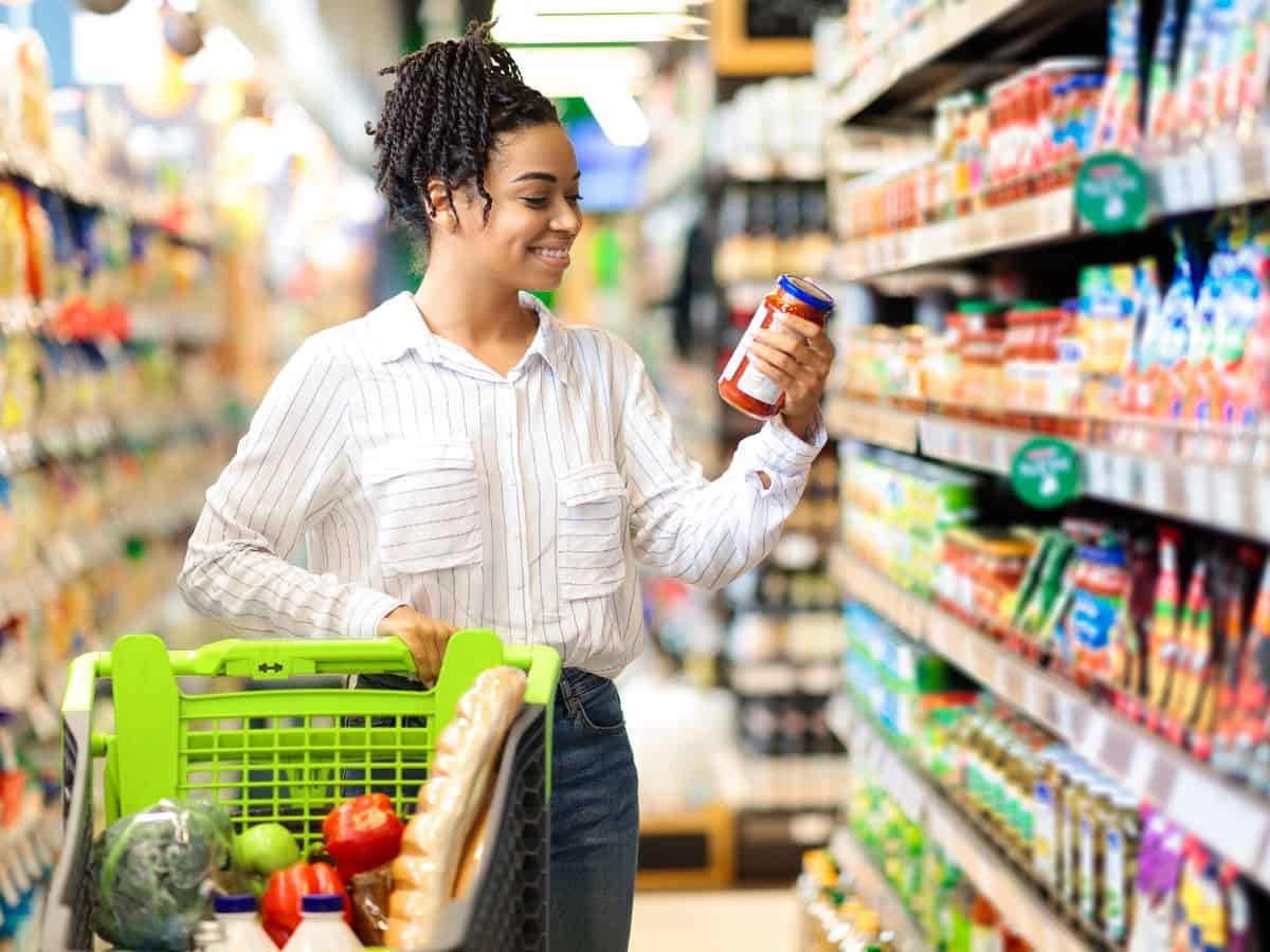 A woman joyfully examines a jar in a grocery aisle filled with colorful food products, representing the comparison of everyday shopping costs, likely hinting at the cost of living differences between Los Angeles vs New York City.
