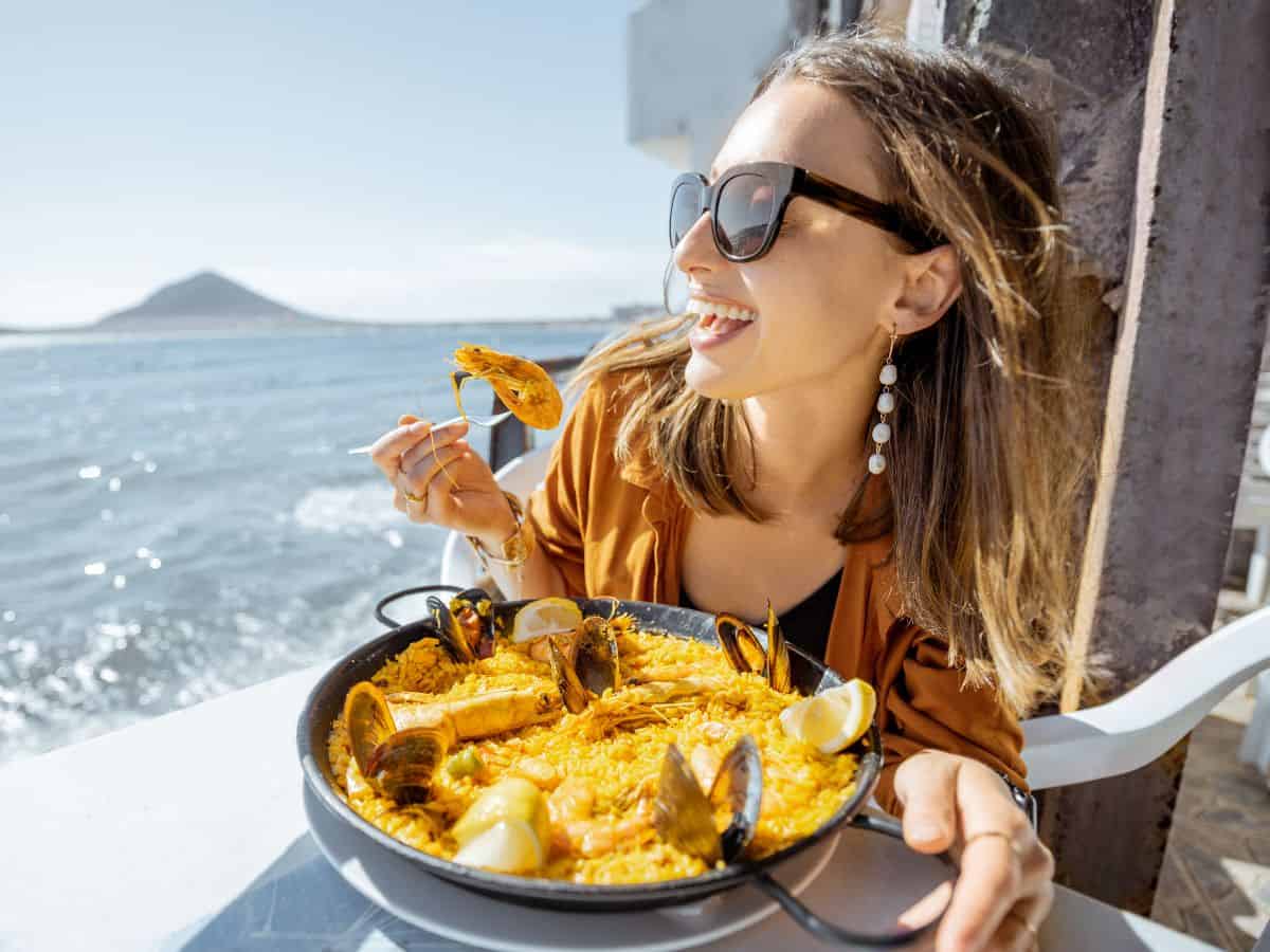 A smiling woman savoring a plate of seafood paella while sitting at an outdoor restaurant overlooking the water. This image captures the food culture in Seattle vs. San Francisco, showcasing seafood cuisine and the joy of dining by the waterfront.