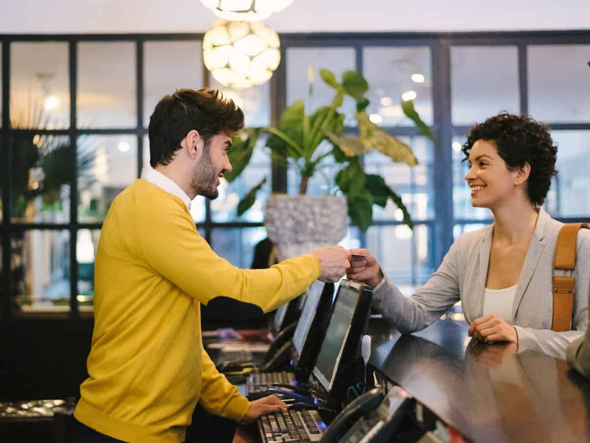 A friendly hotel receptionist in a yellow sweater handing a key card to a smiling guest, highlighting the hospitality and lodging options in the Seattle vs Los Angeles comparison.
