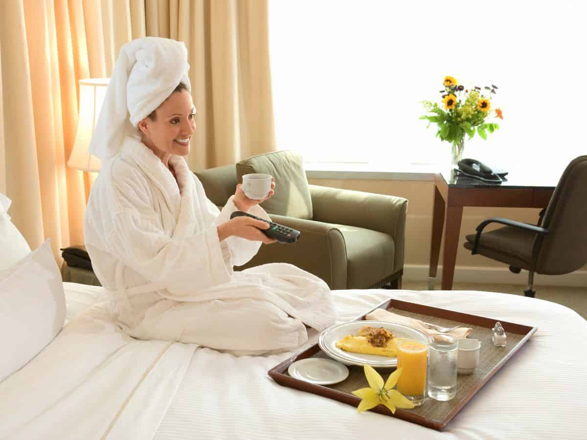  A woman in a bathrobe enjoys breakfast in bed, watching TV, and sipping coffee in a well-lit hotel room. This scene highlights the comfort and relaxation of lodging options that might be considered in either Los Angeles vs New York City.