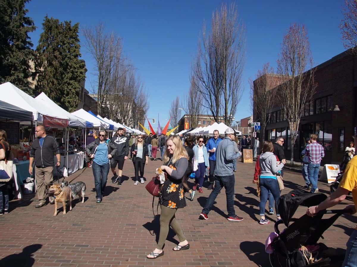 People walking through an open-air farmers market in Seattle on a sunny day, with various stalls set up along a brick-paved street. Shoppers, some with dogs and strollers, browse under white tents. This scene represents the population lifestyle and community culture in Seattle vs. San Francisco.