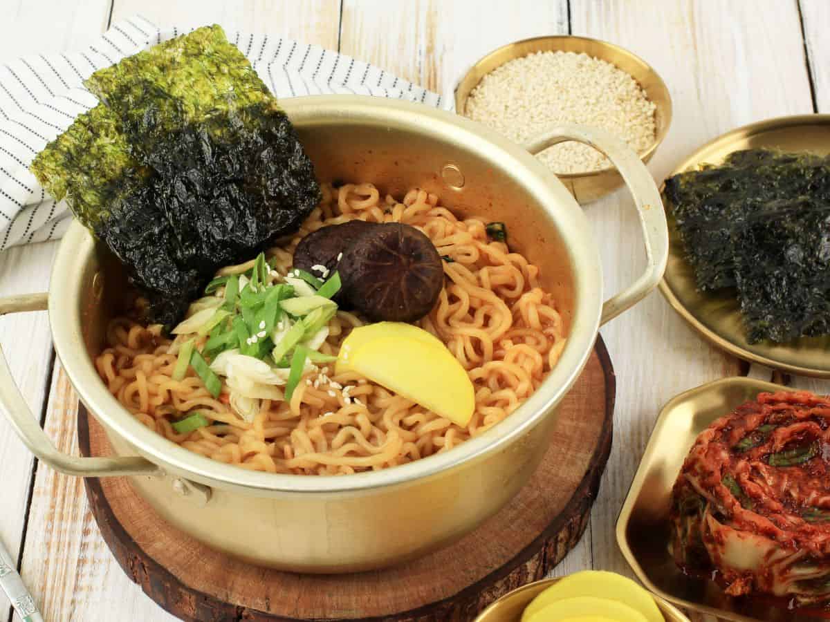 A golden pot of noodles garnished with seaweed, mushrooms, and fresh scallions. The dish is served alongside a plate of kimchi and sesame seeds, embodying the fusion of flavors present in San Francisco's hot pot scene.