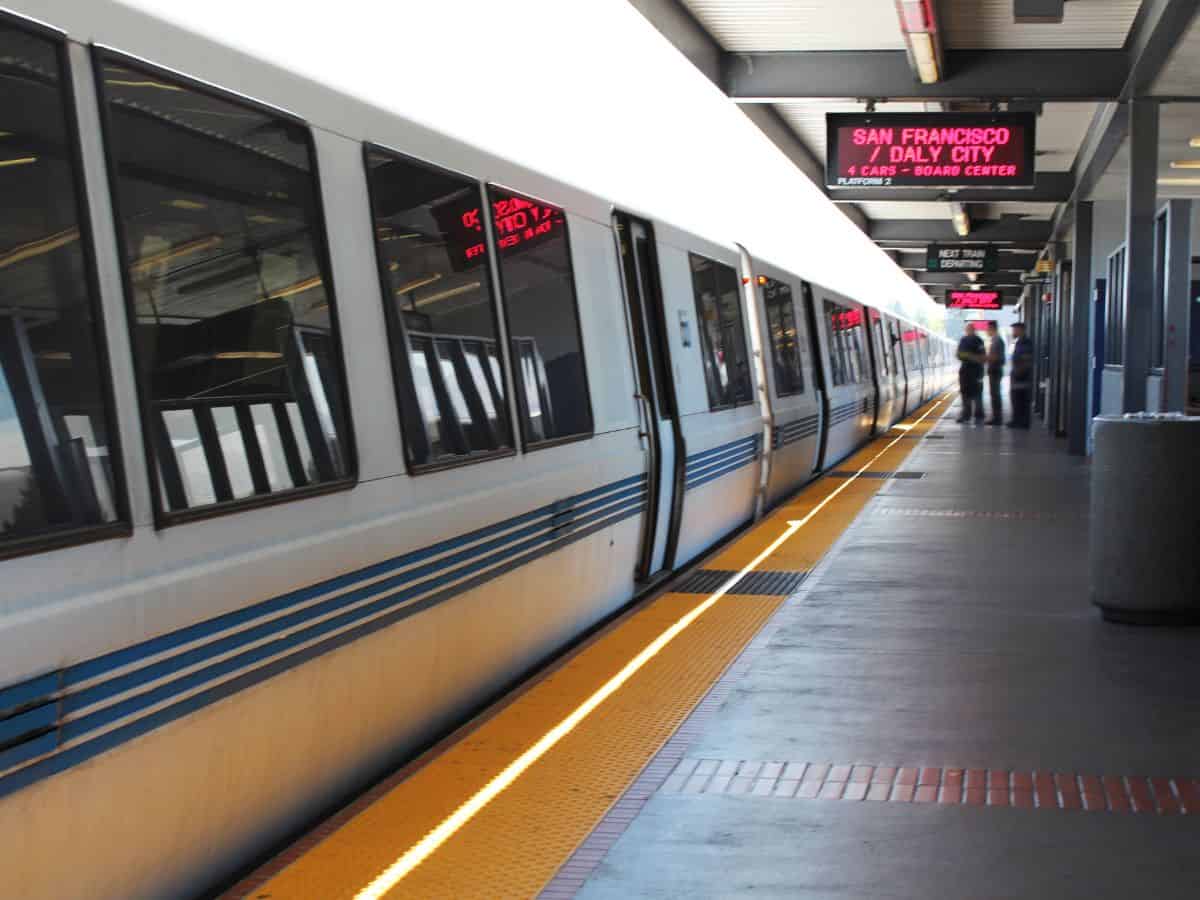 A BART train at a station in San Francisco, with people waiting on the platform. The train is silver with blue stripes, representing public transportation systems in Seattle vs. San Francisco and emphasizing ease of travel and urban mobility.