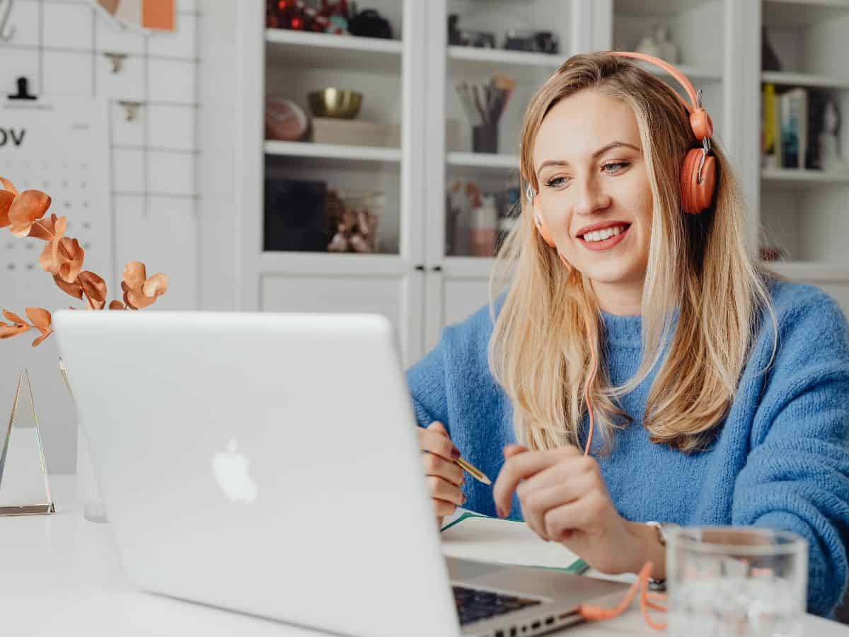 A smiling woman working remotely at a desk with headphones on, in a cozy home office setup. The rise of remote work in the film industry makes Hollywood less reliant on a physical location, contributing to the trend of Hollywood leaving California.
